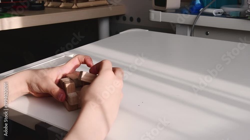 Hands assembling a wooden 3D brain teaser puzzle on a white desk in bright sunlight. Concept of logic, education, and problem solving