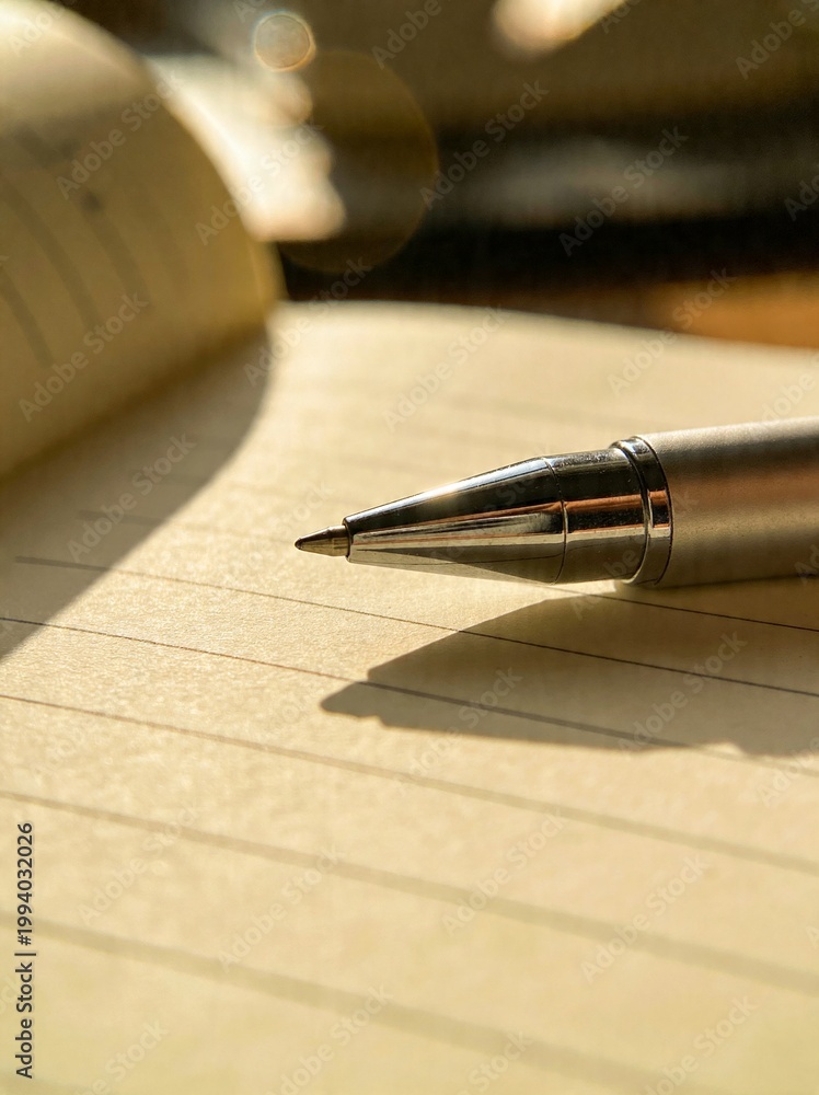Fototapeta premium Close up of a silver ballpoint pen resting on a lined notebook with warm sunlight
