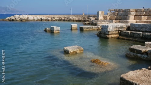 A serene harbor scene with weathered stone blocks protruding from calm waters near a historic dock on a clear day.
