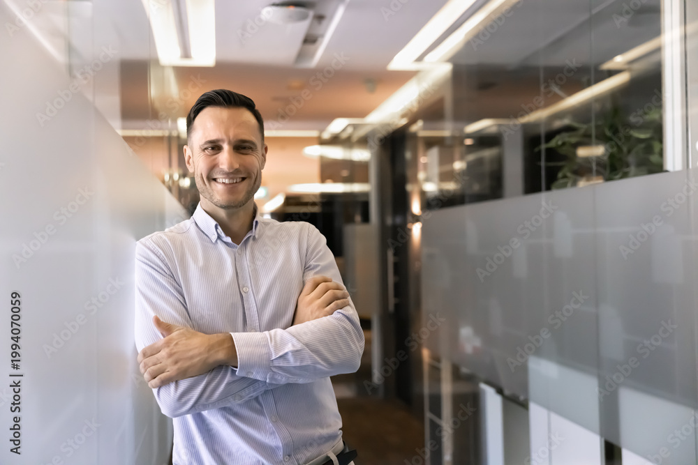 Fototapeta premium Portrait of confident business man or employee stands with arms crossed in office corridor, smiling, looking at camera, represents career growth and professional achievements. Corporate member profile