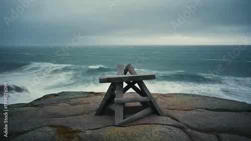 A lone picnic table sits on a rocky cliffside overlooking turbulent ocean waves on a stormy day with gloomy skies.