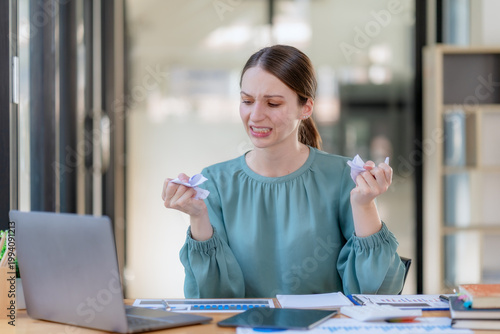 Angry businesswoman tearing up paper document at office desk with frustrated expression.