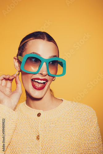 Happy woman with large blue sunglasses and red lipstick smiles brightly against yellow background. She wears a textured beige sweater and gold earrings. Concept of fashion and joy.