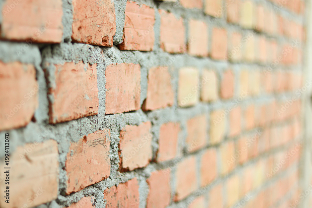 Fototapeta premium Side view of an unfinished red brick wall with selective focus. Rough clay bricks and grey mortar texture. Industrial background for construction or architecture projects.