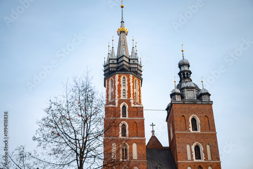 Close-up of the two asymmetrical brick towers of St. Mary's Basilica in Krakow against a pale sky. Detailed gothic architecture featuring ornate spires and golden crowns.