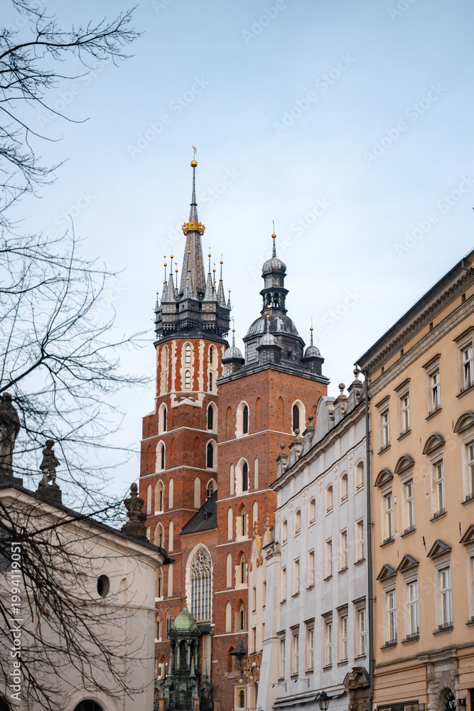Obraz premium Vertical view of the iconic Gothic towers of St. Mary's Basilica rising above the historic architecture of Krakow's Old Town square under a clear, soft morning sky in Poland.