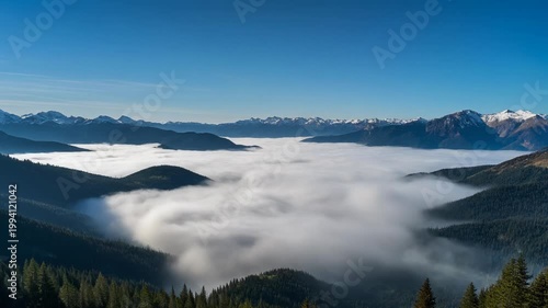 A serene mountain landscape with fog rolling over hills and trees under a clear blue sky with distant snowy peaks