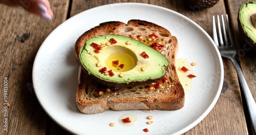 Hand placing sliced avocado on toasted bread with chili flakes and olive oil on a rustic table