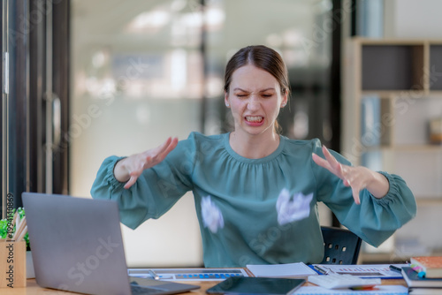 Angry businesswoman tearing up paper document at office desk with frustrated expression.