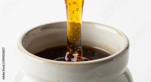 Thick golden honey is slowly poured from a dripping utensil into a ceramic pot filled with dark liquid.