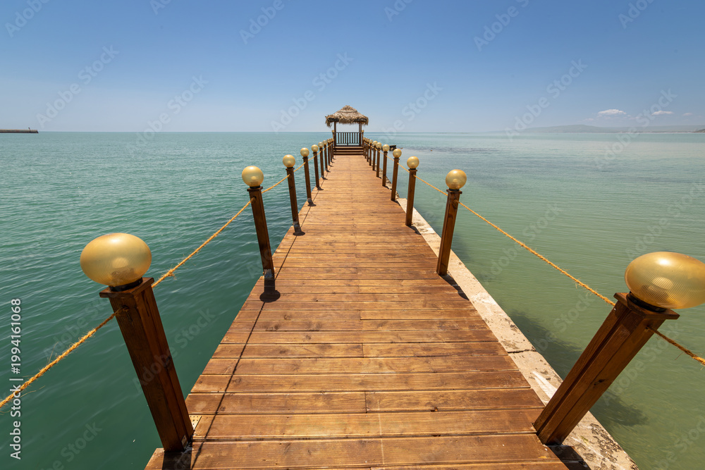 Fototapeta premium Idyllic wooden pier with a thatched gazebo stretching into the calm turquoise sea under a clear blue sky. Serene summer vacation background, perfect for travel, relaxation, and tourism concepts.