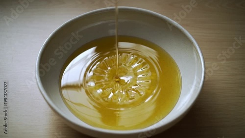 A white bowl with honey pouring into it, containing a lemon slice on a wooden table with warm tones.