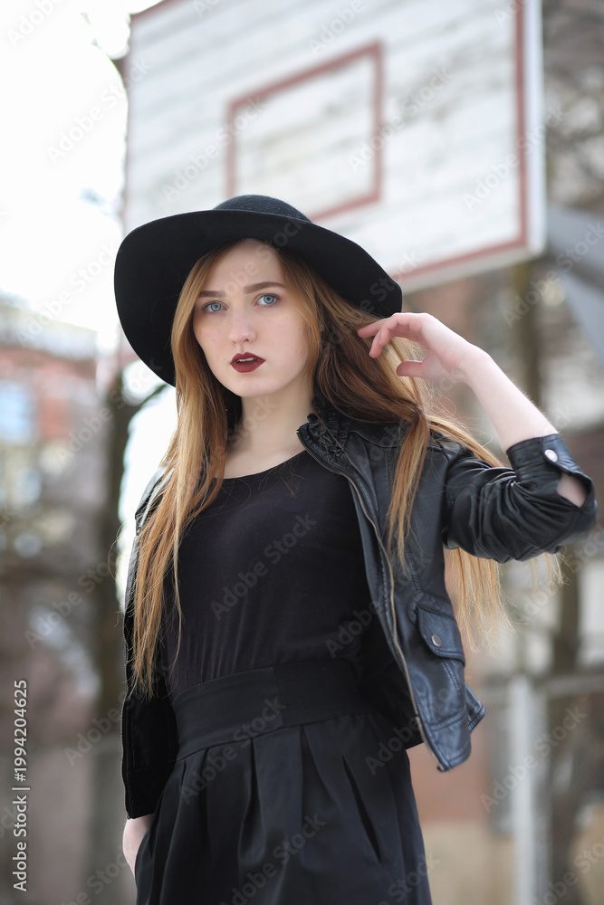 Fototapeta premium A girl walks down the city street in a leather waistcoat with a phone. Young beautiful girl in a hat and with a dark make-up outside. Girl in the Gothic style on street.