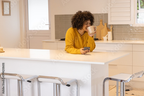 African American woman in mustard knit leaning on white kitchen island, sipping mug, checking phone