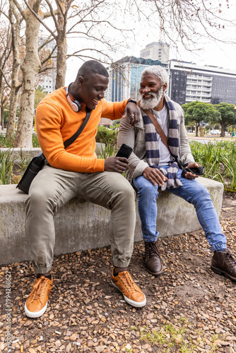 African American male friends, senior sitting on bench at park holding phones in orange sweatshirt