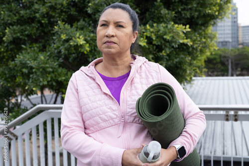 Senior Hispanic woman standing on metal rail stairs in pink jacket holding green mat, clear bottle