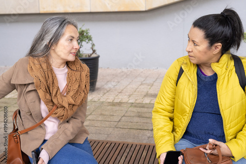Mature women chatting on wood bench in plaza wearing beige coat and yellow puffer, holding bags