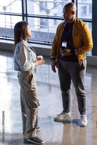 Diverse coworkers talking and holding takeaway coffee cups in atrium wearing blue lanyard badge