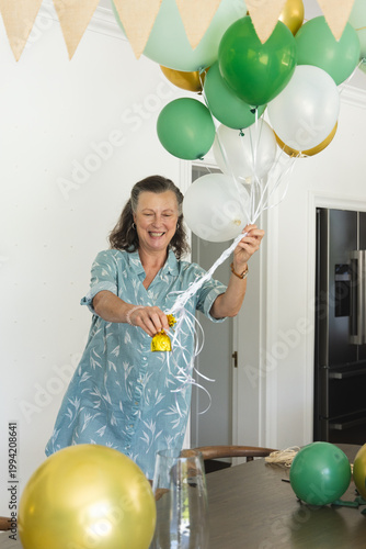Senior woman standing at table in kitchen wearing blue dress holding green white gold balloons