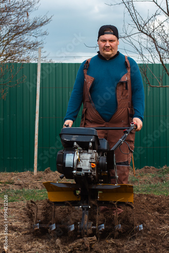 Farmer in work clothes operating a walk-behind tractor, cultivating and preparing the brown soil of a home garden or field for planting crops in a rural outdoor setting.
