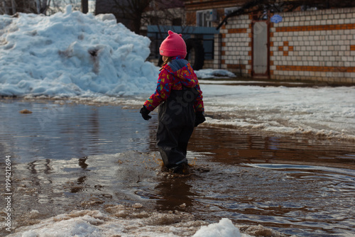 Child wearing winter clothes and a pink hat playing in a large muddy puddle, surrounded by melting snow and slush, enjoying a playful outdoor activity during a sunny winter day.