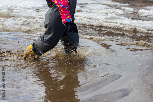 Child wearing waterproof clothing and boots jumping into a large muddy puddle, creating splashes and enjoying outdoor activity on a wet day with melting snow visible in the background.