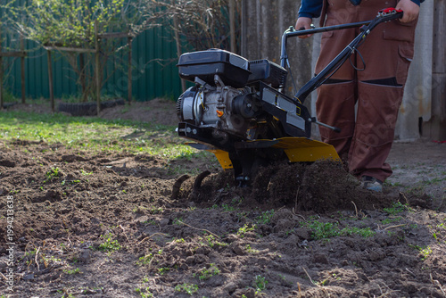 Man operating a walk-behind tractor to till and turn brown soil in a backyard garden plot, preparing fertile ground for spring planting and small-scale farming tasks.