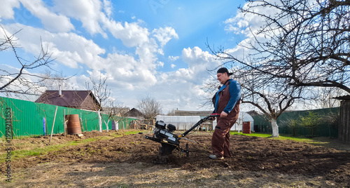 Farmer or gardener preparing agricultural land for planting by tilling earth with a motor tiller, breaking up soil clumps in a rural vegetable garden on a sunny spring day.