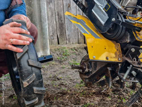 Person's hands preparing to attach a new tire to the axle of a yellow walk-behind tractor, performing essential maintenance for spring farming and cultivation work.