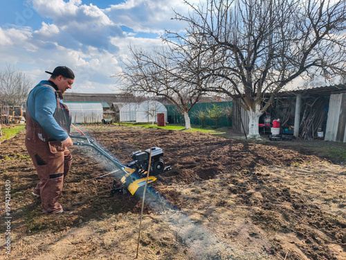Man cultivating his home garden plot during springtime, preparing the ground for planting using a rototiller to aerate and loosen the soil for agricultural work.