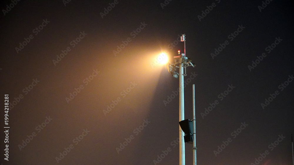 Obraz premium High mast airport lighting pole against dark misty night sky illumination for aviation ground operations and terminal safety security transportation infrastructure background