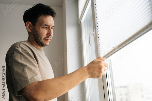 Slika na platnu Thoughtful man with worried expression opens white pleated blinds looking outside