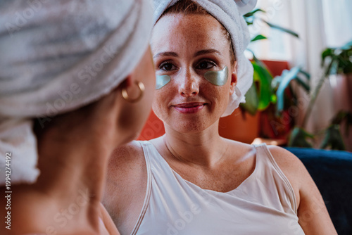 Two women wearing towels on their heads and applying eye patches for beauty treatment