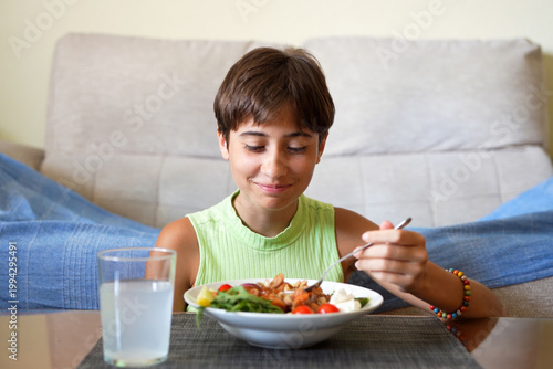 Smiling child relishing nutritious homemade salad with glass of milk nearby in bright living room