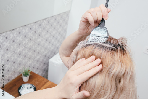 Woman applying white hair dye to roots of blonde hair with brush at home in bathroom