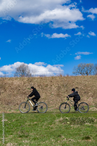 Two cyclists enjoy a sunny day riding mountain bikes across a grassy field under a vibrant blue sky.