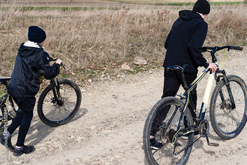 Two kids walking their bikes down a dirt road. A sunny day outdoors.
