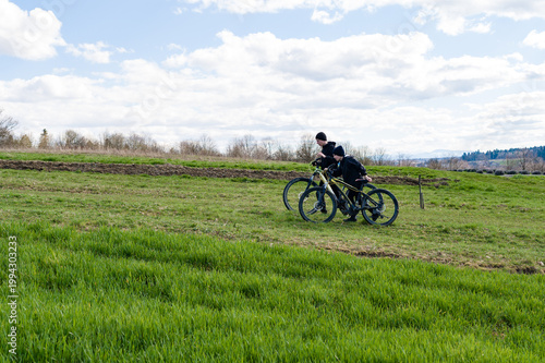 Two cyclists pause on a grassy field, enjoying the sunny landscape.
