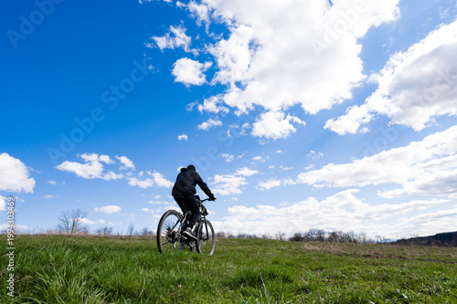 Person cycling uphill under a vibrant blue sky dotted with fluffy clouds. A peaceful outdoor scene.
