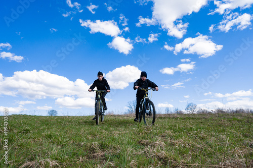 Two boys on mountain bikes enjoying a sunny day outdoors, riding across a grassy field under a bright blue sky.