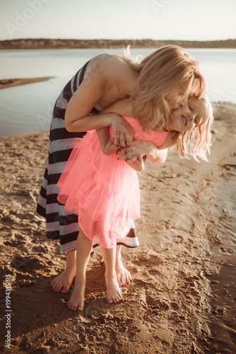 Tableau sur toile Mother kissing her daughter on cheek on sandy shore near calm water during golden hour