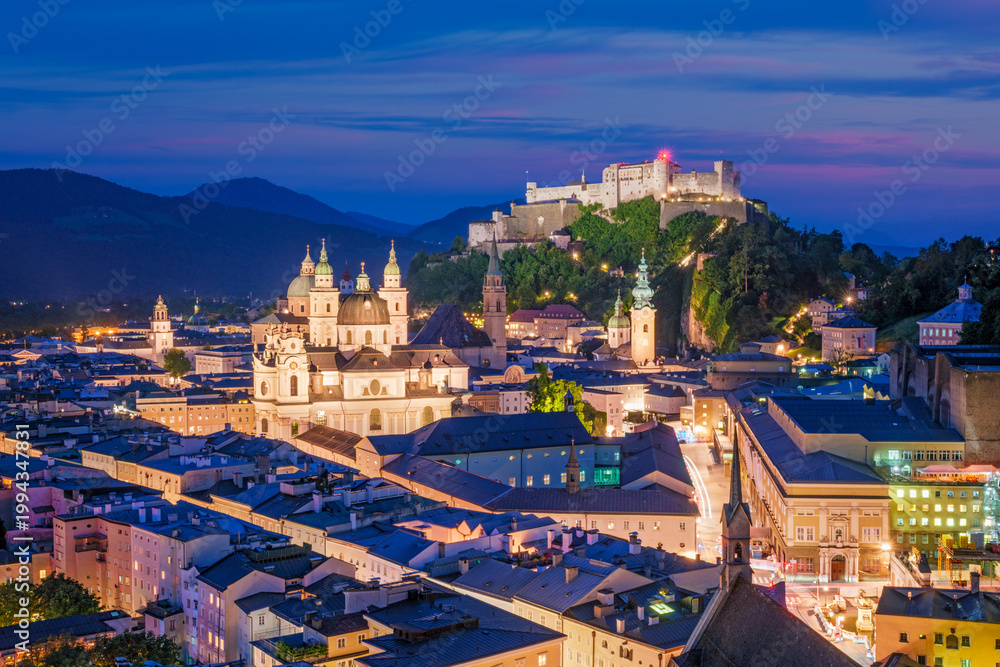 Obraz premium Close-up aerial view of the lit-up Hohensalzburg Fortress and the Salzburg Cathedral during the blue hour in the historic city center of Austria.