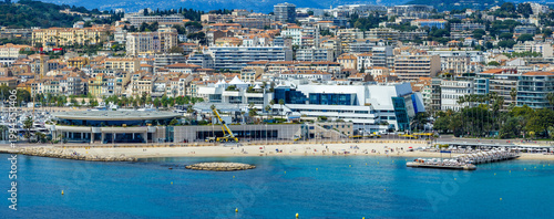 A bright aerial view shows Cannes, France, with the Bay of Cannes, Palais des Festivals, Promenade de la Croisette, sandy beaches, marina, and the Carlton hotel at midday.