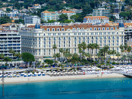 Aerial Cannes, France, shows Carlton Hotel facing turquoise water and sandy beach, palm lined promenade, umbrellas, and loungers in bright daylight during festival season.