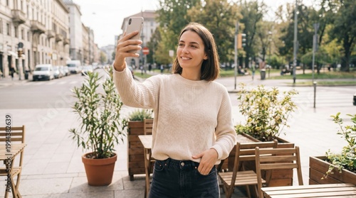 A cheerful young woman in a beige sweater is taking a selfie with a smartphone at an outdoor urban cafe in the city.