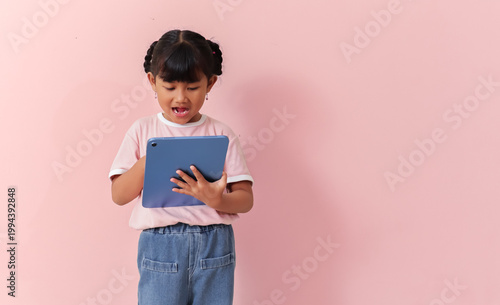 cute young southeast asian girl holding tablet and looking at screen with surprised expression on pink studio background, modern digital lifestyle concept