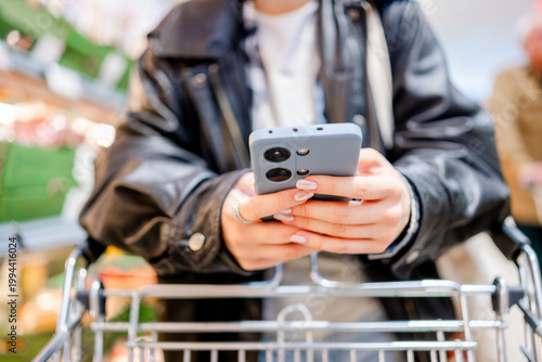 Fotografija Young Girl In Supermarket With Shopping List On Smartphone Goes With A Cart Clos