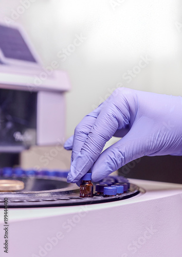 Gloved scientist’s hand placing a biochemical sample tube in the rotary carousel of a high-tech automatic analysis machine