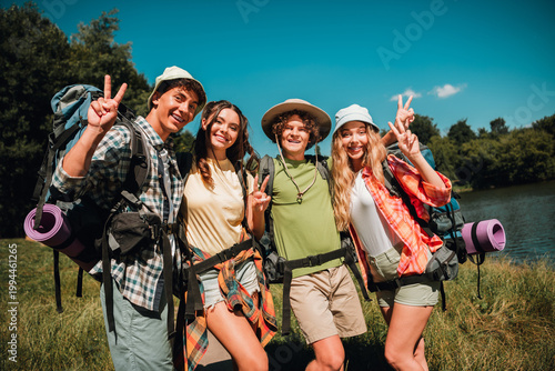 Bild auf Leinwand Four friends on a summer backpacking trip by a lake making peace signs and enjoy