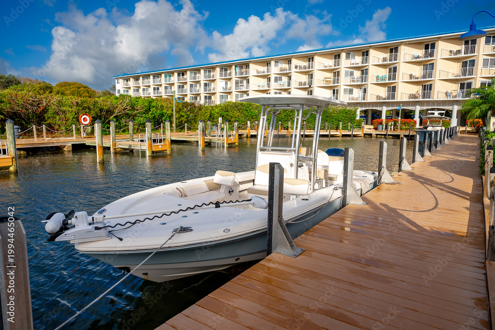 Obraz premium White motorboat docked at a wooden pier near resort building. High angle view of a powerboat moored at a boardwalk in a tropical marina canal.